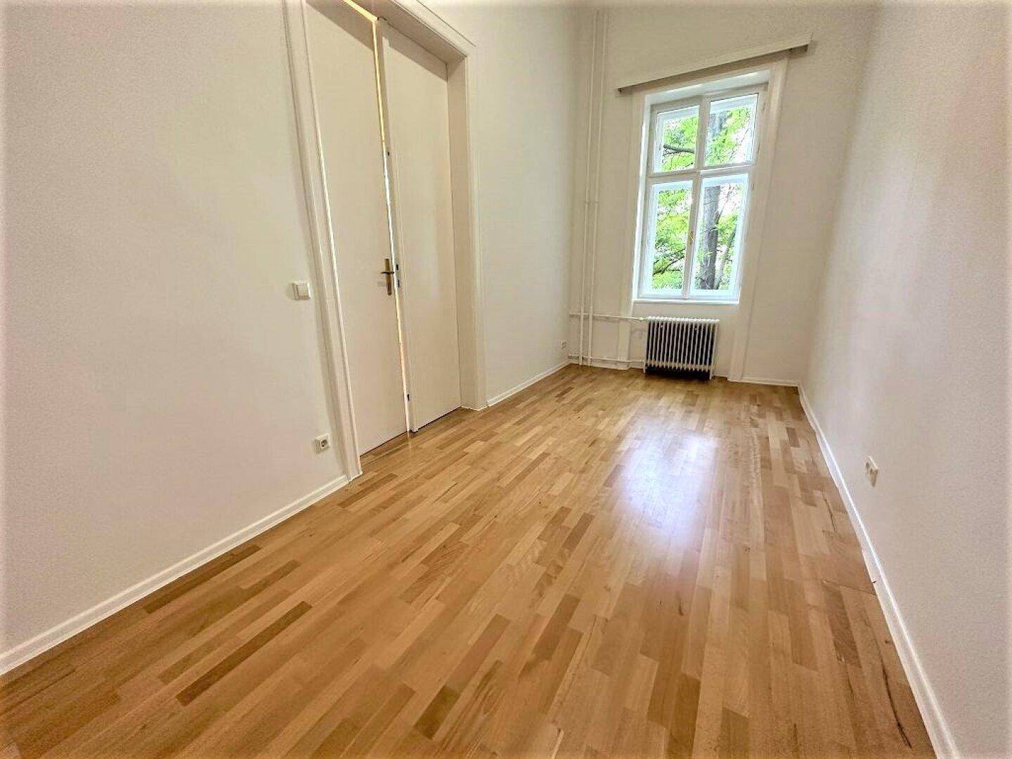 Geräumiges Schlafzimmer mit hellem Parkettboden, hoher Decke und einer weißen Tür.