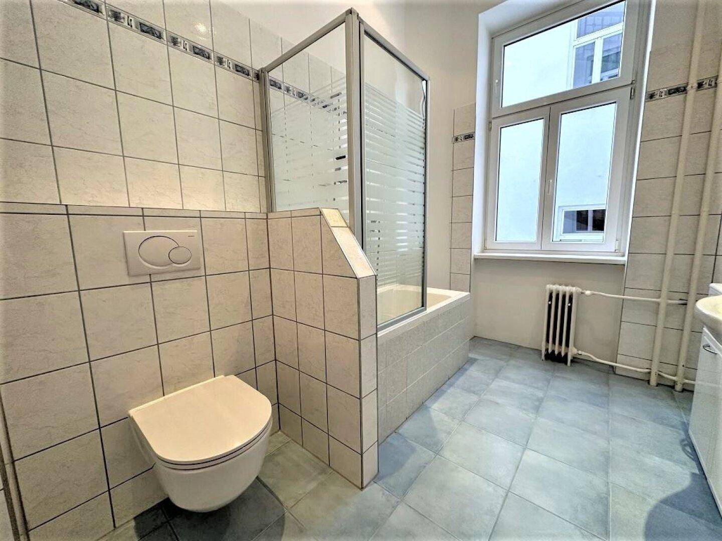 Badezimmer mit Badewanne, Duschabtrennung, WC und Fenster, gefliest in Weiß und Blau.