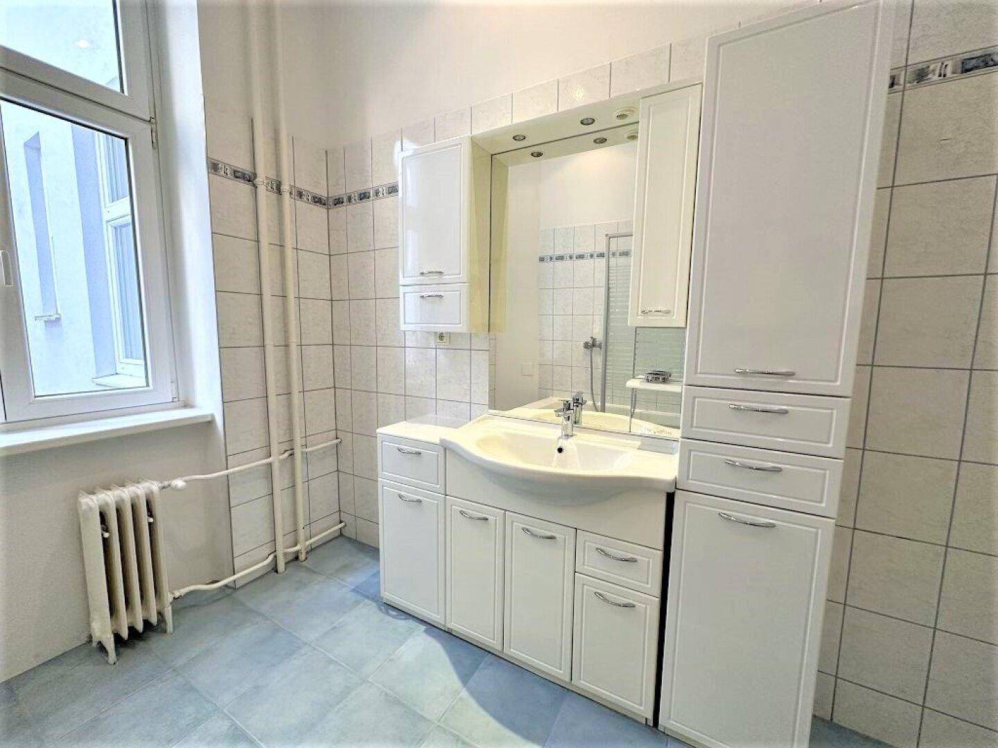 Helles Badezimmer mit großem Waschtisch, Spiegelschrank und Fenster, gefliest in Weiß und Blau.