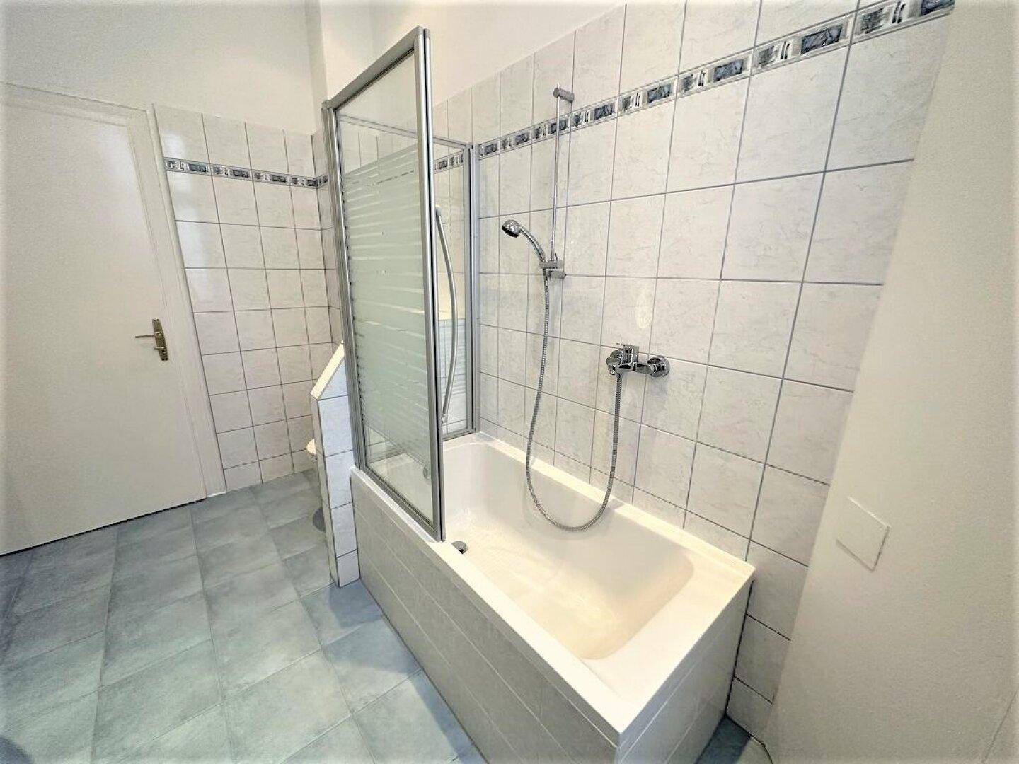 Badezimmer mit Badewanne, Duschabtrennung und weißen Fliesen, kombiniert mit blauen Bodenfliesen.