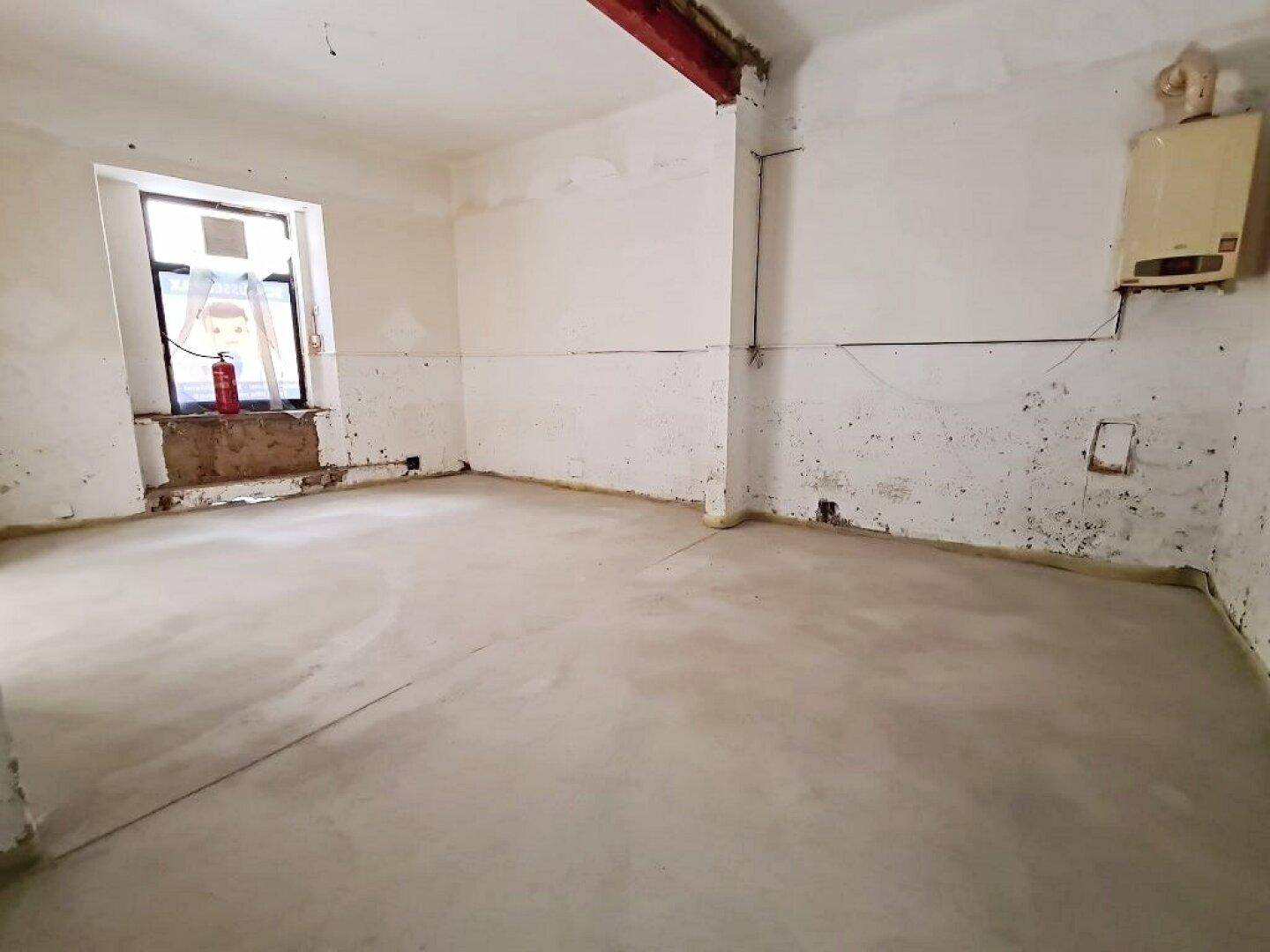 Renovierungsbedürftiger Innenraum mit Betonboden, freigelegten Wänden und einem alten Warmwasserbereiter.