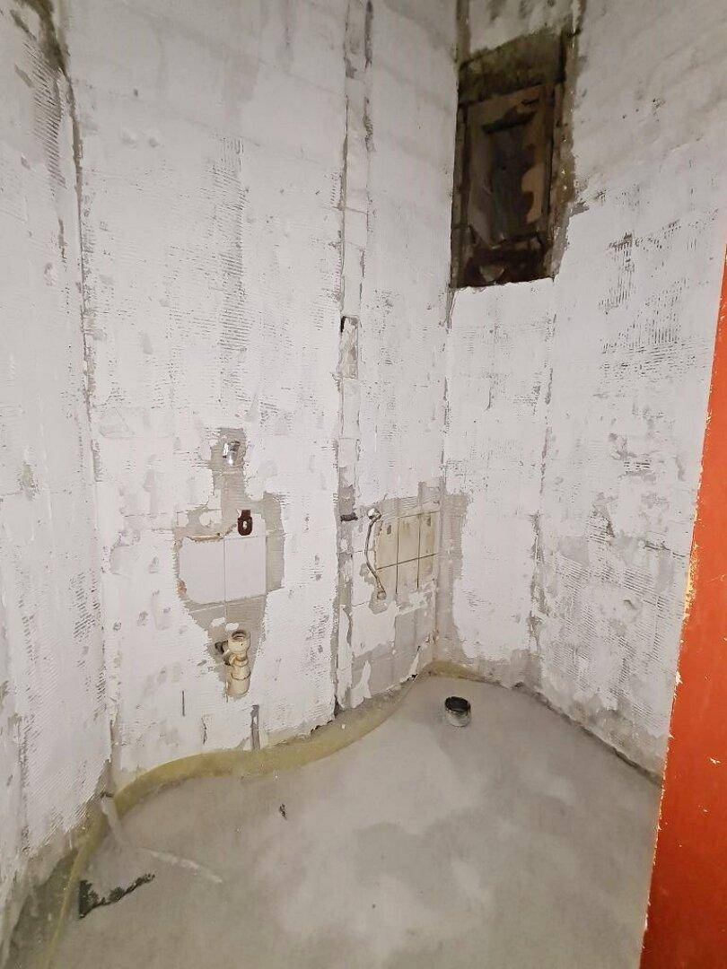 Badezimmer im Rohbauzustand mit freigelegten Wänden, sichtbaren Rohrleitungen und einem kleinen Fenster.