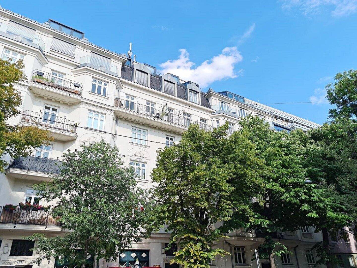 Klassische Fassade eines Mehrfamilienhauses mit Balkonen und grünen Bäumen im Vordergrund unter blauem Himmel.