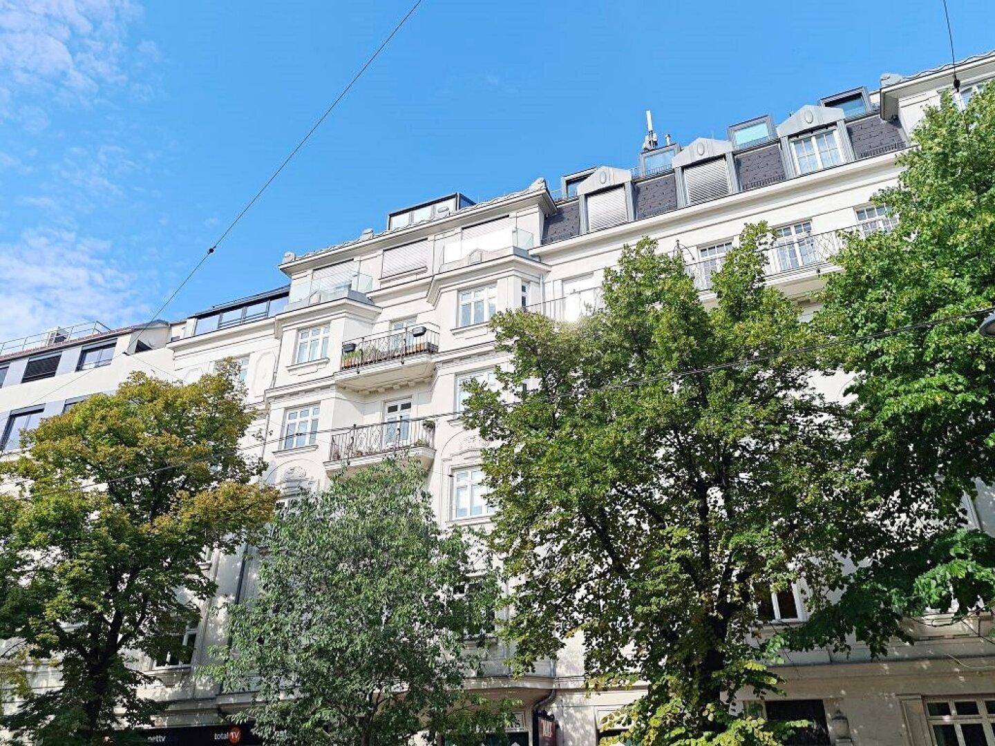 Klassische Fassade eines Altbaus mit mehreren Balkonen und grünen Bäumen davor unter blauem Himmel.