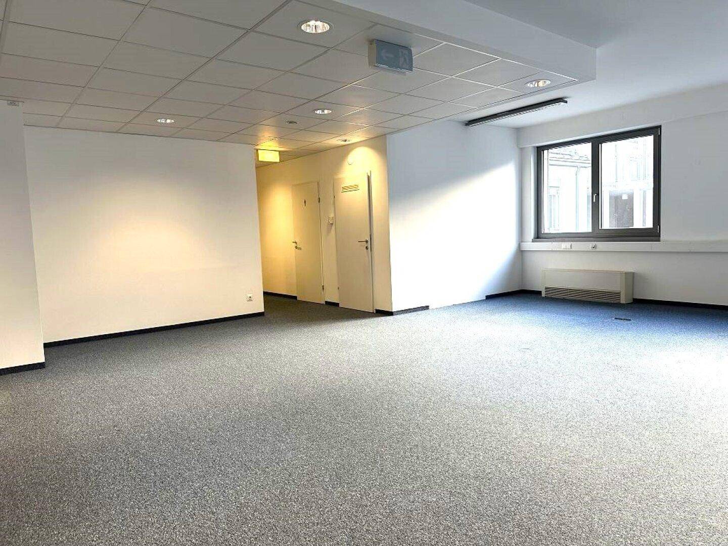 Großer, heller Büroraum mit Teppichboden und Fenstern, die viel Tageslicht hereinlassen.