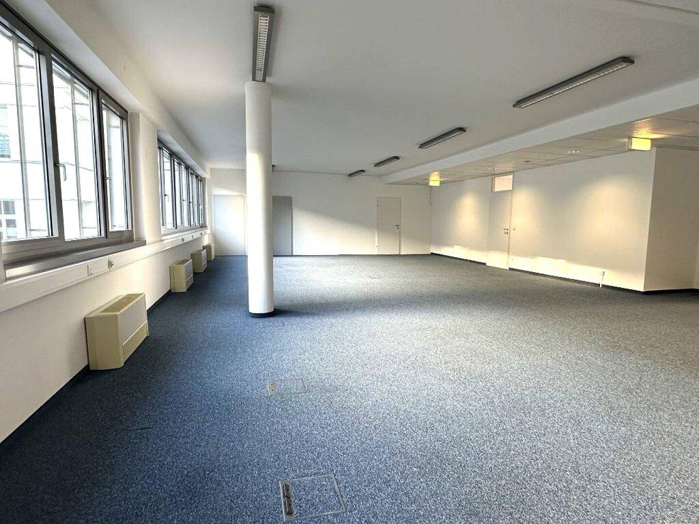 Geräumiger Büroraum mit Teppichboden, Säule und großen Fenstern, die viel Tageslicht hereinlassen.