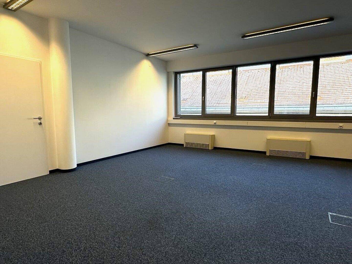 Heller Büroraum mit Teppichboden und Fenstern, die einen Blick auf die Umgebung bieten.