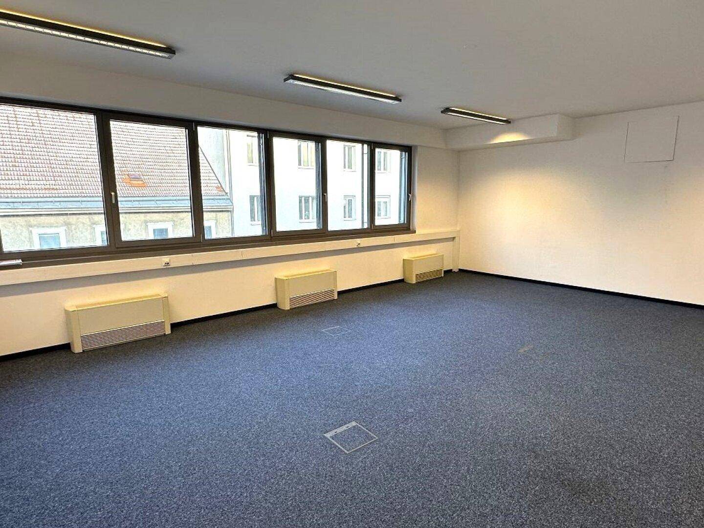 Geräumiger Büroraum mit Teppichboden und großen Fenstern, die viel Tageslicht hereinlassen.