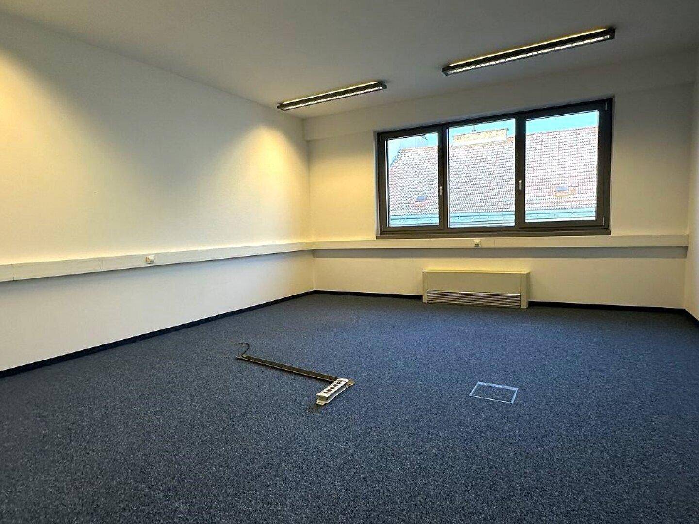 Heller Büroraum mit Teppichboden und Fenstern, die einen Blick auf die Umgebung bieten.