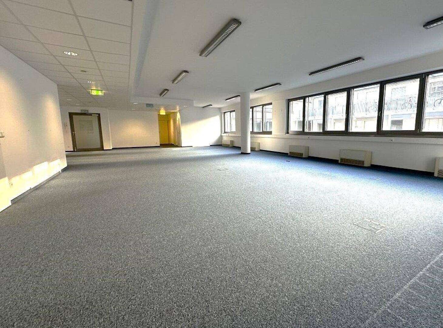 Großer, leerer Büroraum mit Teppichboden und vielen Fenstern, ideal für flexible Arbeitsplatzgestaltung.
