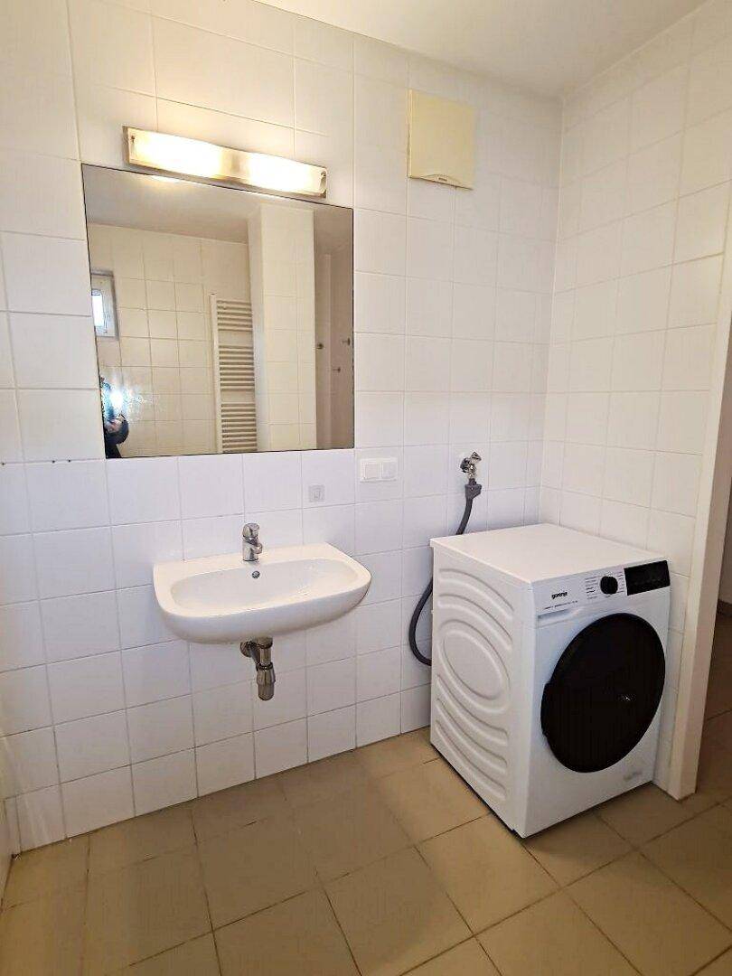 Badezimmer mit Waschbecken, Spiegel und Platz für eine Waschmaschine, hell gefliest.
