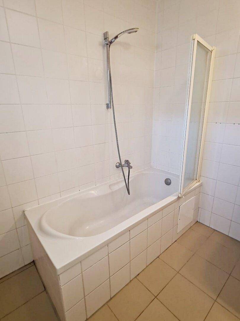 Einfache Badewanne mit Duscharmatur und Duschwand in einem hell gefliesten Badezimmer.