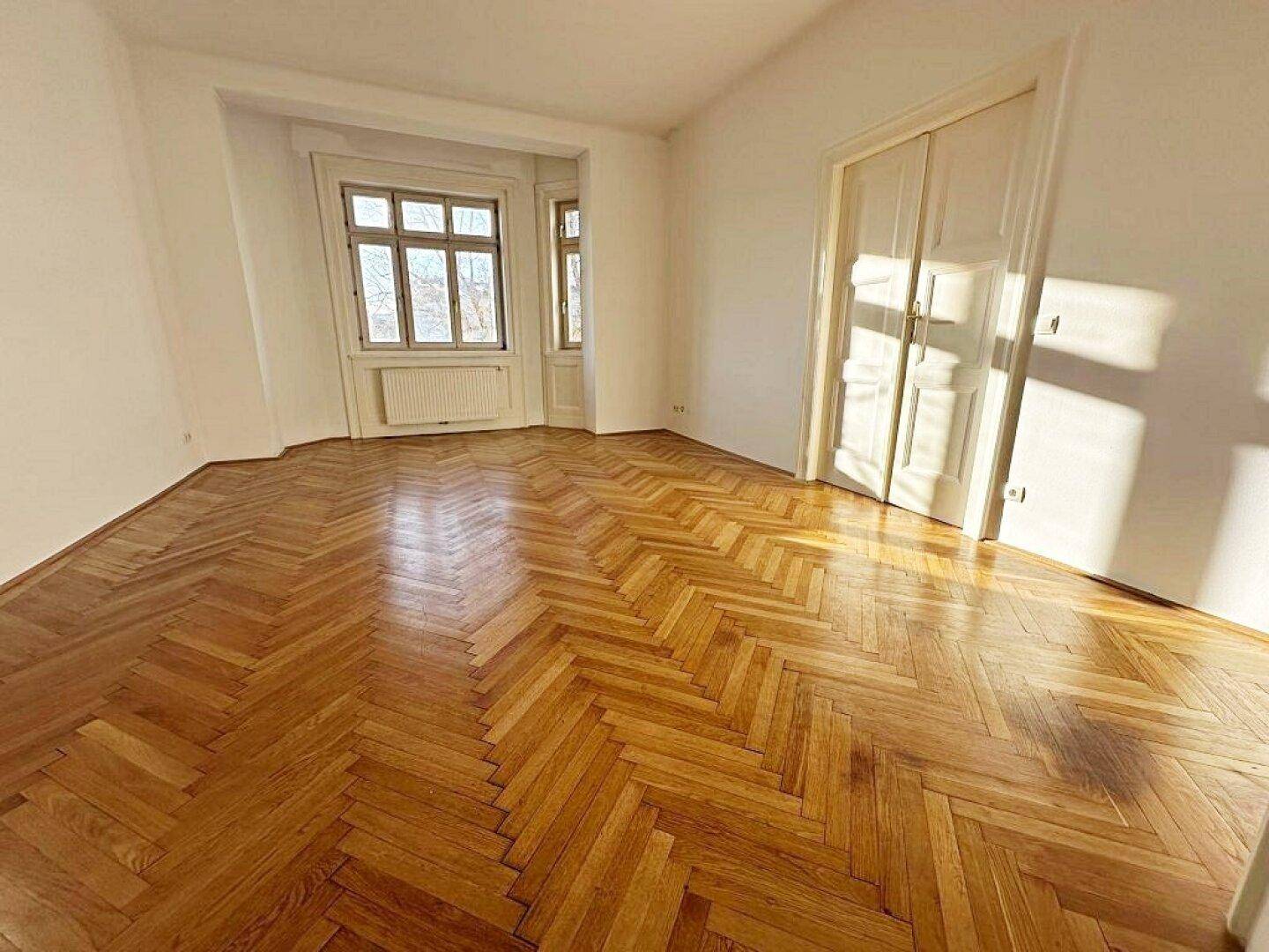 Helles Zimmer mit Fischgrätparkettboden, weißen Wänden und einem Fenster, das viel Sonnenlicht hereinlässt.