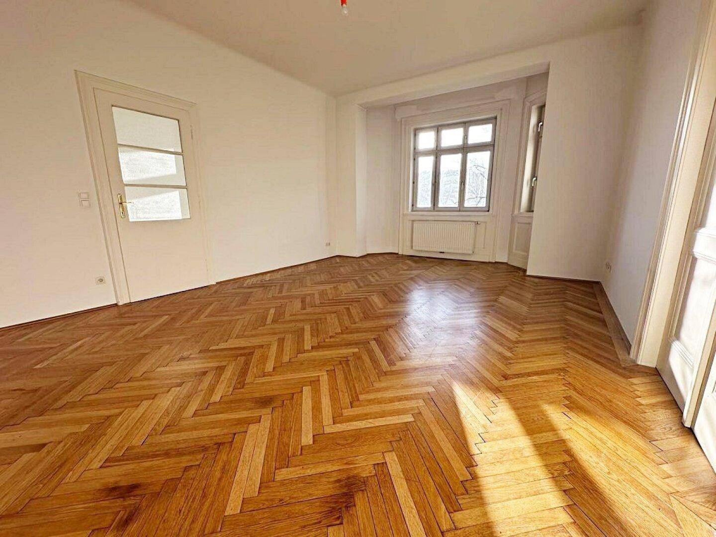 Geräumiges Zimmer mit glänzendem Fischgrätparkett, weißen Wänden und einem großen Fenster für viel Tageslicht.