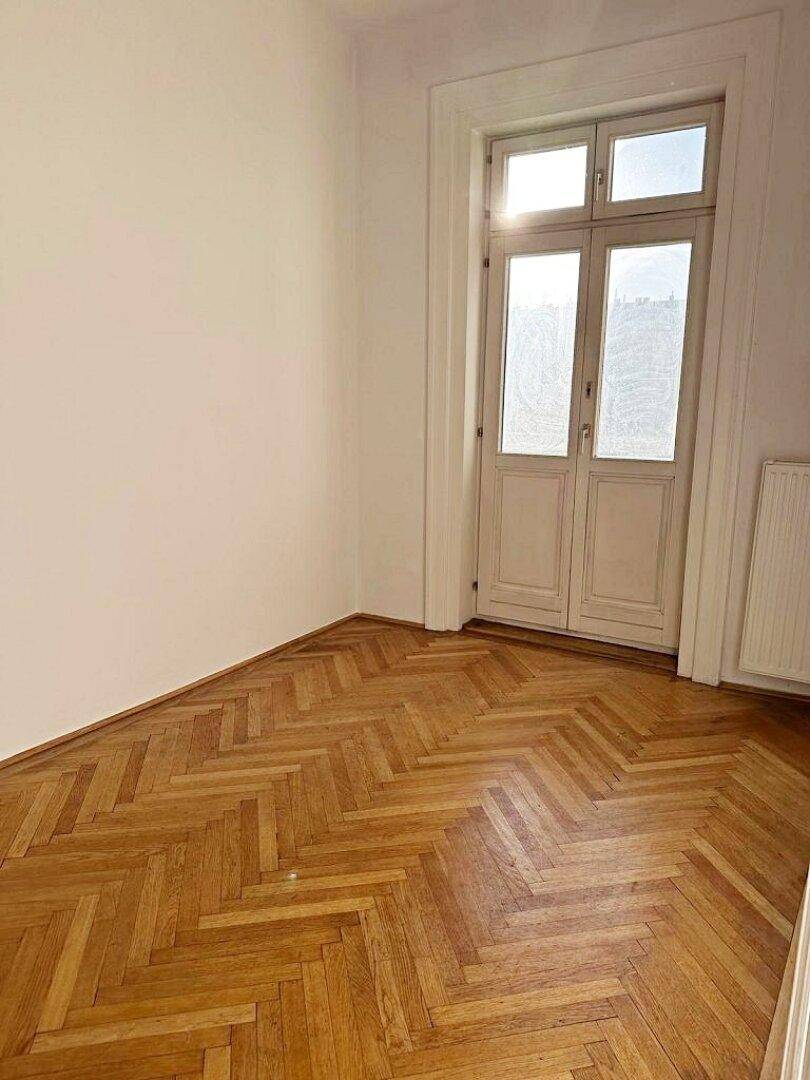 Lichtdurchflutetes Zimmer mit Fischgrätparkett und einer hohen Fenstertür, die nach draußen führt.