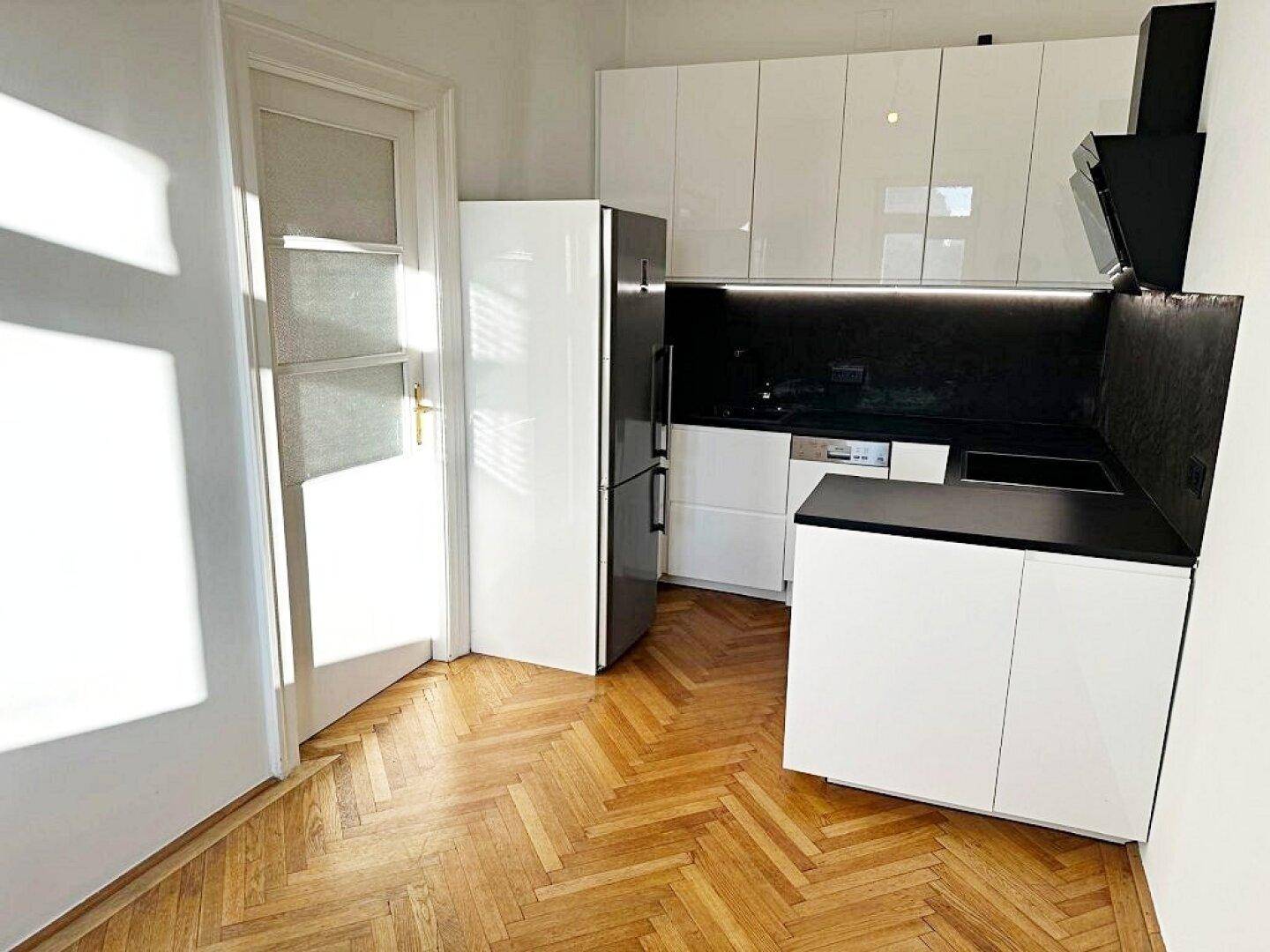 Moderne Küche mit weißen Hochglanzschränken, schwarzen Arbeitsplatten und Fischgrätparkettboden.