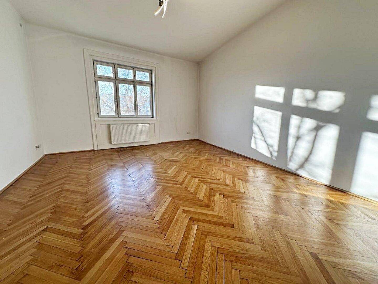 Helles Zimmer mit Fischgrätparkettboden, weißen Wänden und einem Fenster, das viel Sonnenlicht hereinlässt.