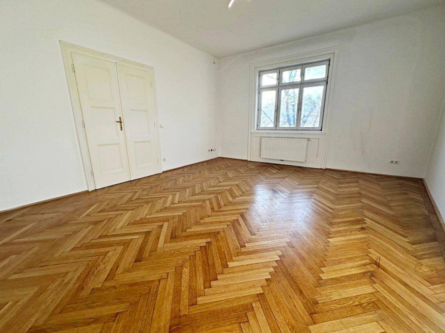Geräumiges Zimmer mit Fischgrätparkett, weißen Wänden und einem Fenster mit Heizkörper darunter.