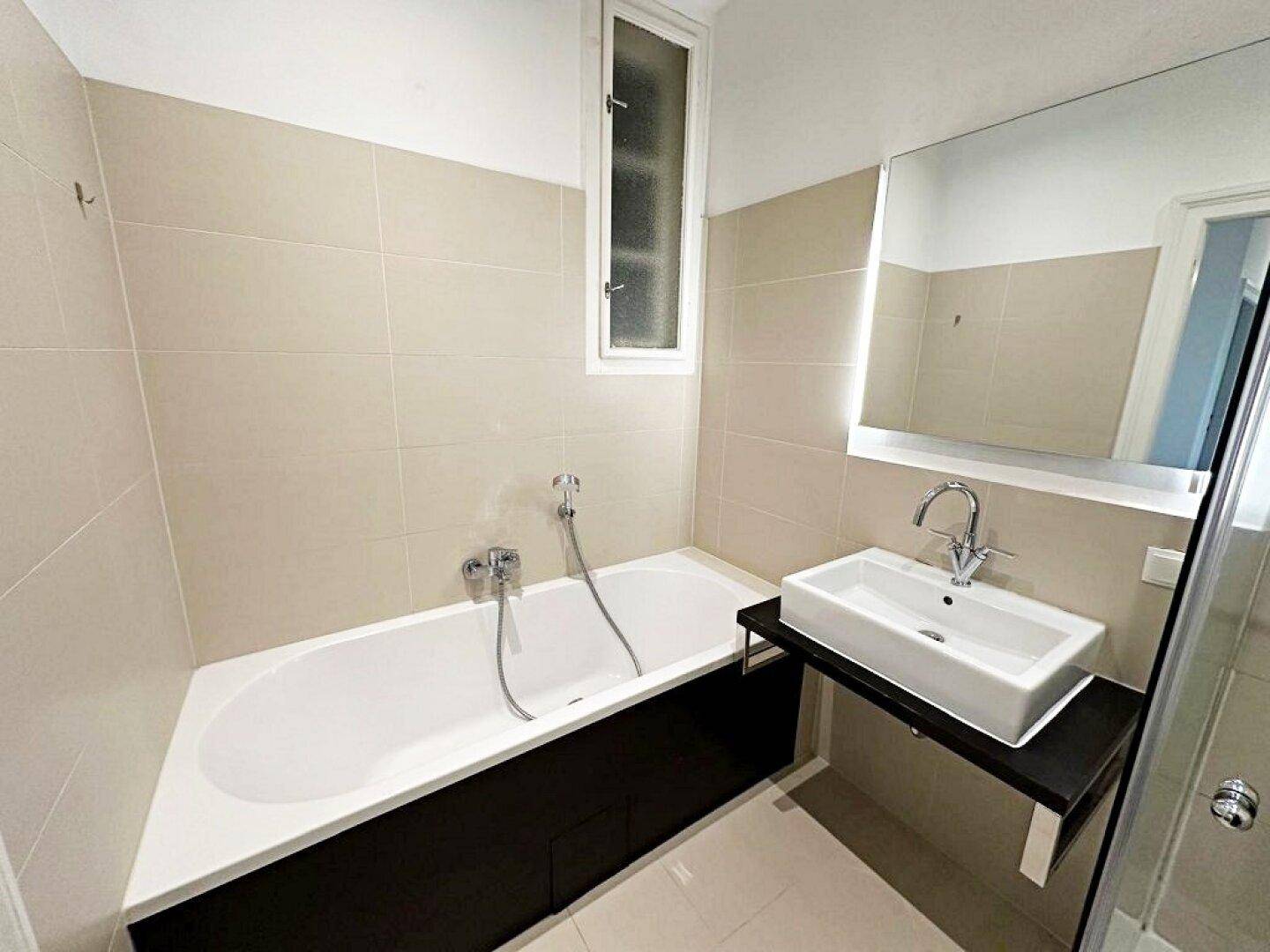 Modernes Badezimmer mit Badewanne, Waschbecken und beigefarbenen Fliesen, gut beleuchtet durch ein schmales Fenster.
