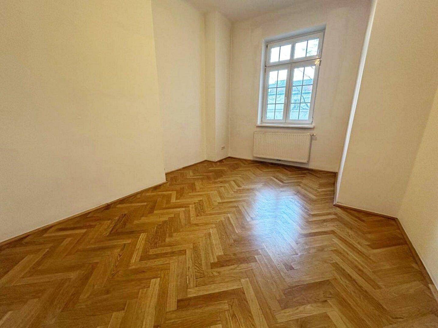 Kleineres Zimmer mit Fischgrätparkett, hellen Wänden und einem Fenster mit Heizkörper.