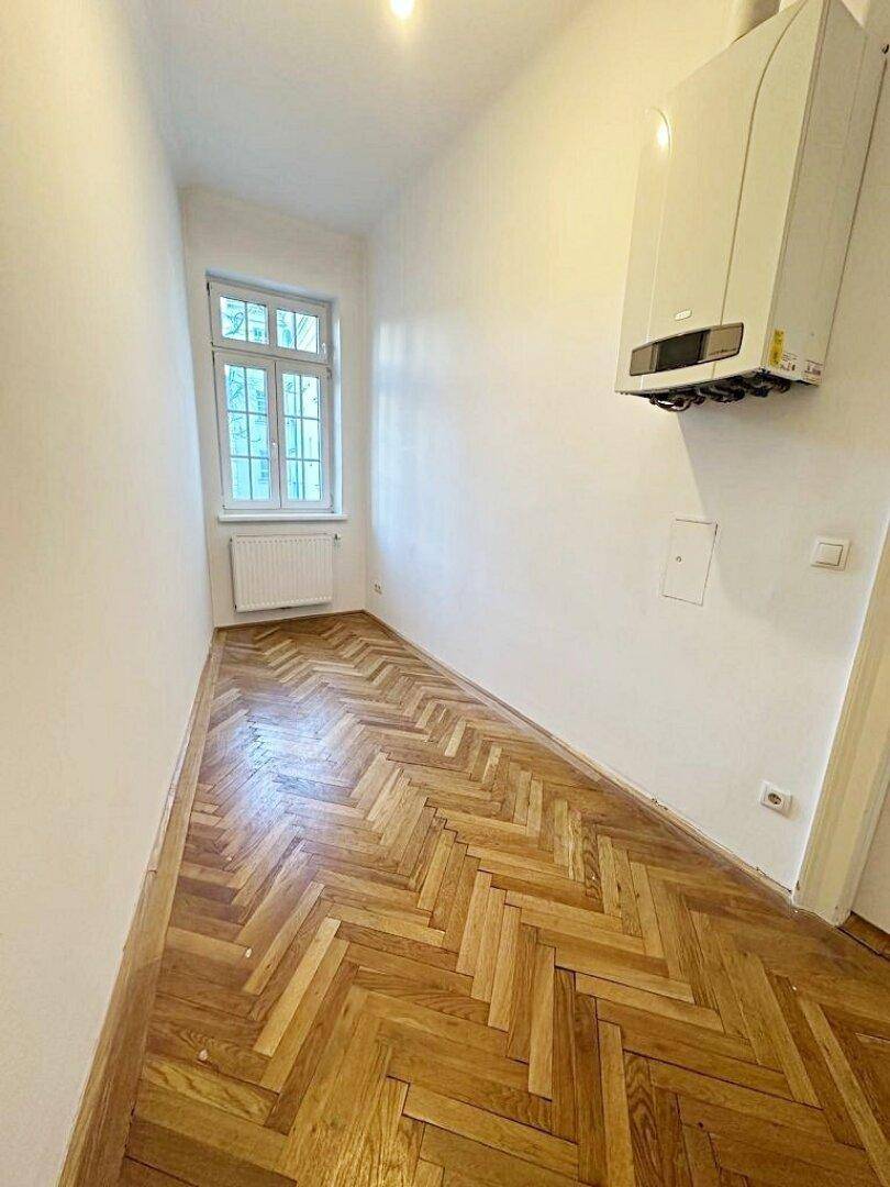 Schmaler Flur mit Fischgrätparkett, Fenster und einem wandmontierten Heizkessel.