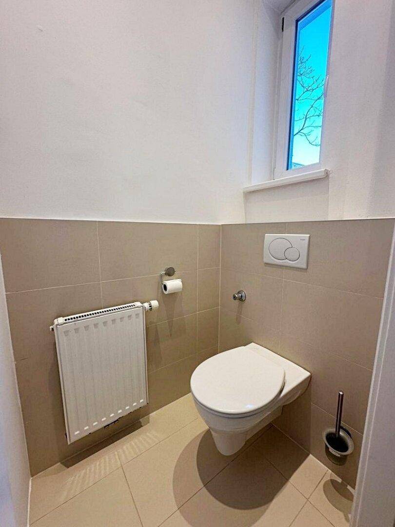 Separates WC mit wandhängender Toilette, Heizkörper und einem hohen Fenster für natürliches Licht.