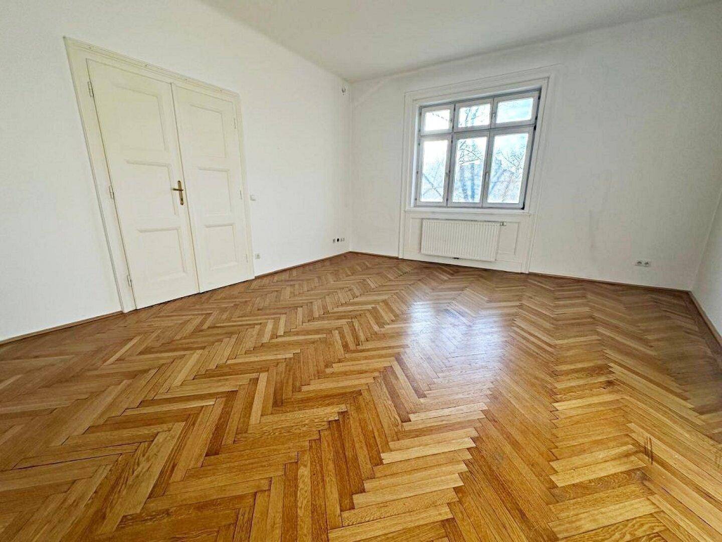 Geräumiger, heller Raum mit glänzendem Fischgrätparkett und großem Fenster, ideal als Wohn- oder Schlafzimmer.