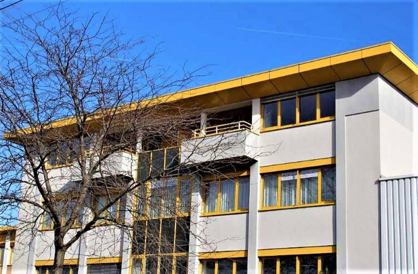 Seitenansicht des Gebäudes mit gelben Fensterrahmen und einem Balkon unter blauem Himmel.