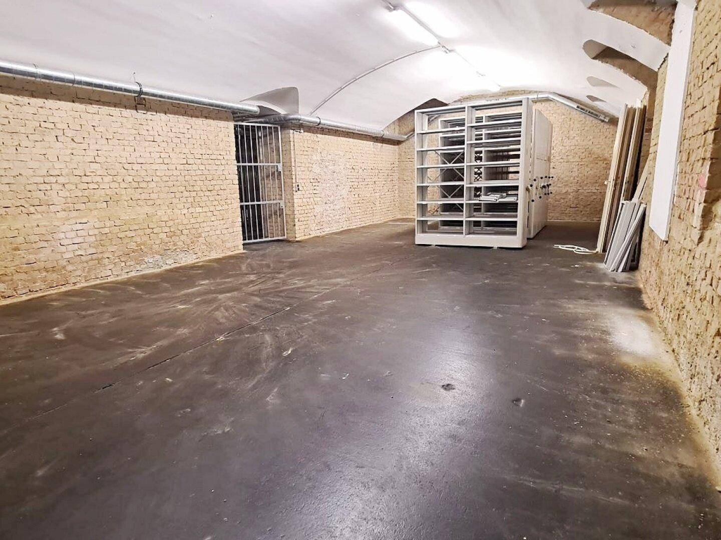Kellerraum mit unverputzten Ziegelwänden, Betonboden und einem robusten Metallgittertor.