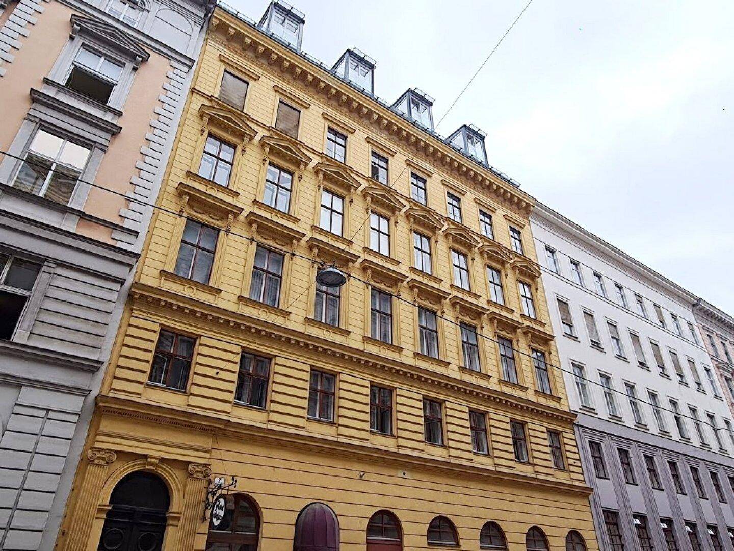 Historisches Gebäude mit gelber Fassade, vielen Fenstern und dekorativen Elementen in einer städtischen Umgebung.
