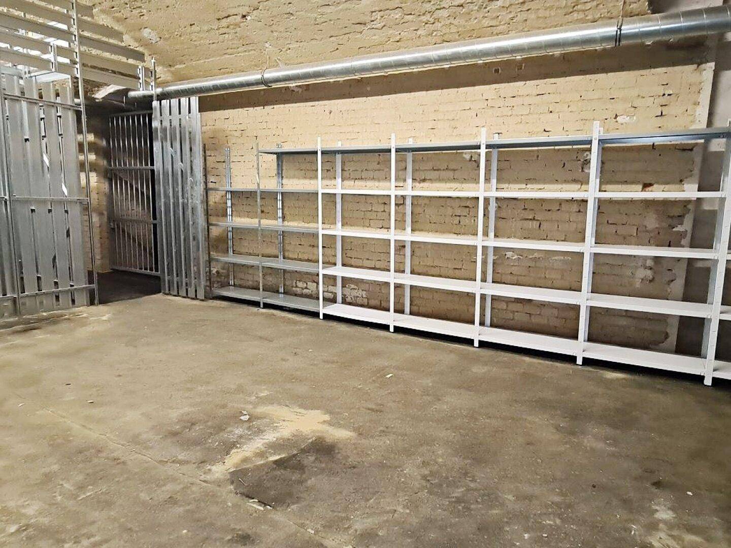 Langer Kellerraum mit durchgehenden Metallregalen an der Ziegelwand und stabilen Metallabtrennungen im Vordergrund.