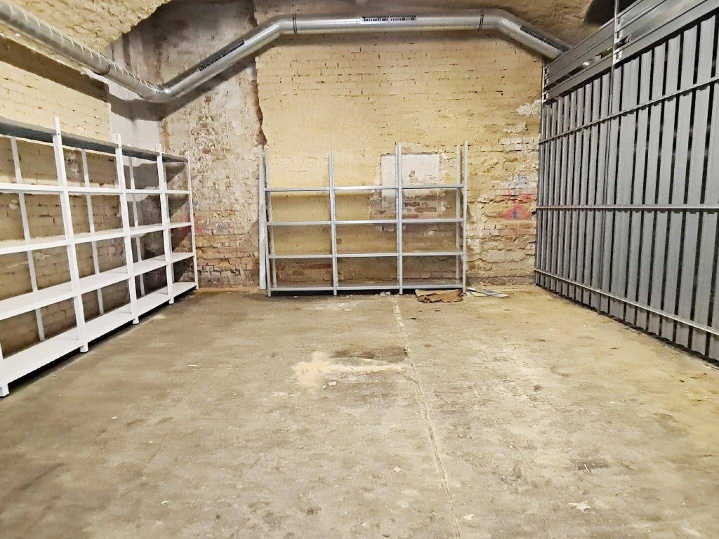Kellerbereich mit Metallregalen an der Ziegelwand, Betonboden und sichtbaren Lüftungsrohren unter der gewölbten Decke.