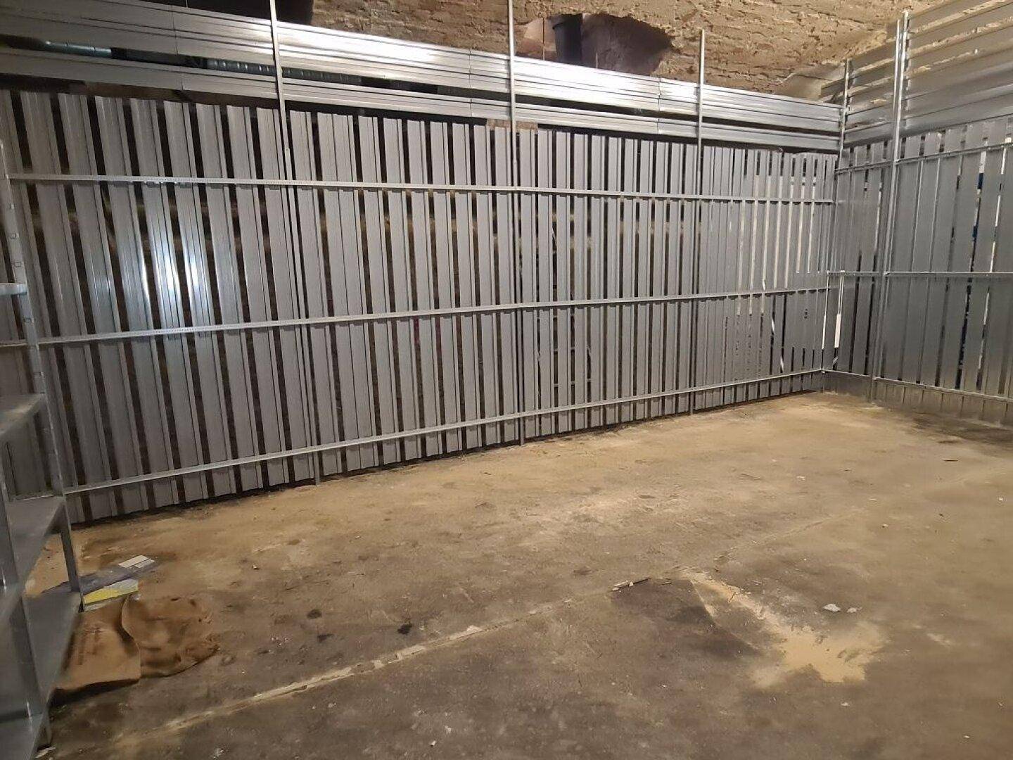 Kellerraum mit Metallabtrennungen, die Lagerbereiche definieren, und einem einfachen Betonboden.