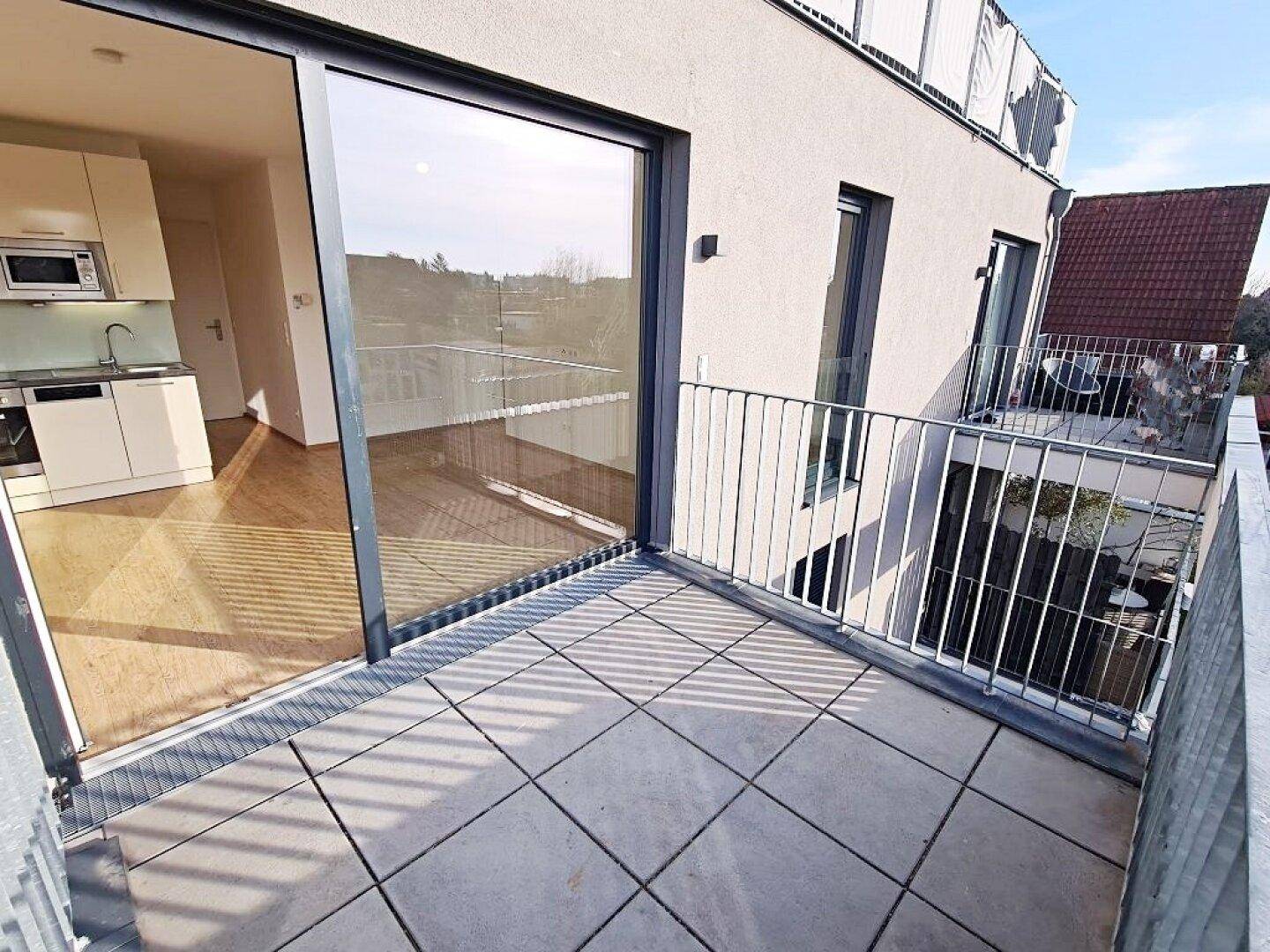 Geräumiger Balkon mit Fliesenboden und Blick in den hellen Innenraum mit offener Küche.