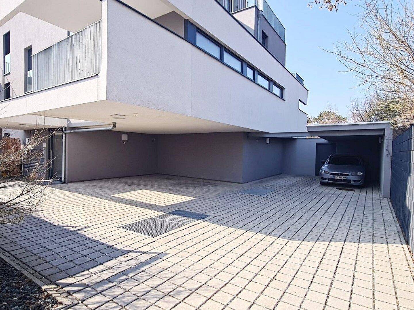 Moderner Eingangsbereich mit gepflastertem Vorplatz und überdachtem Carport für Fahrzeuge.