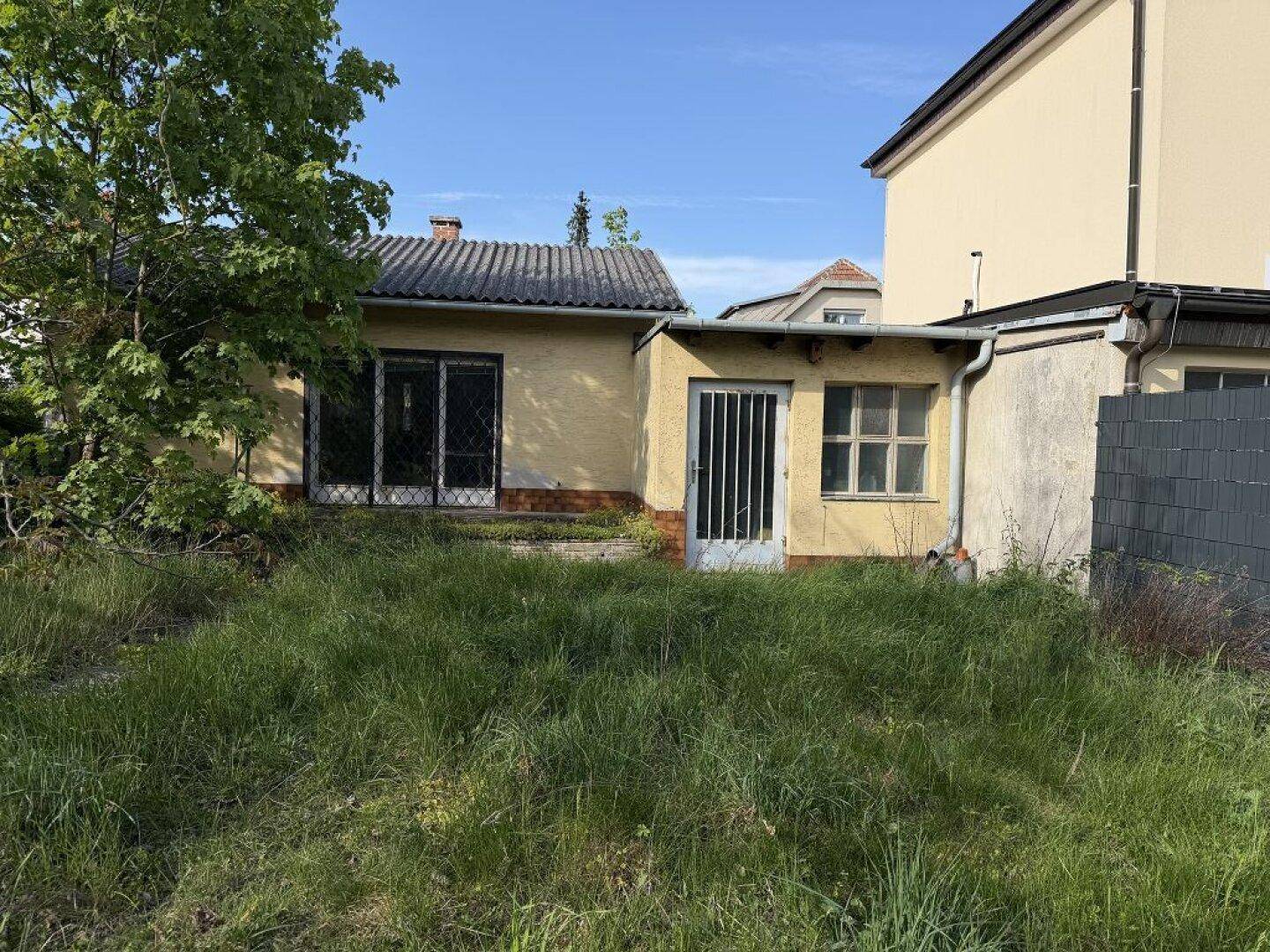 Rückansicht eines älteren Hauses mit verwildertem Garten und Anbau unter blauem Himmel.