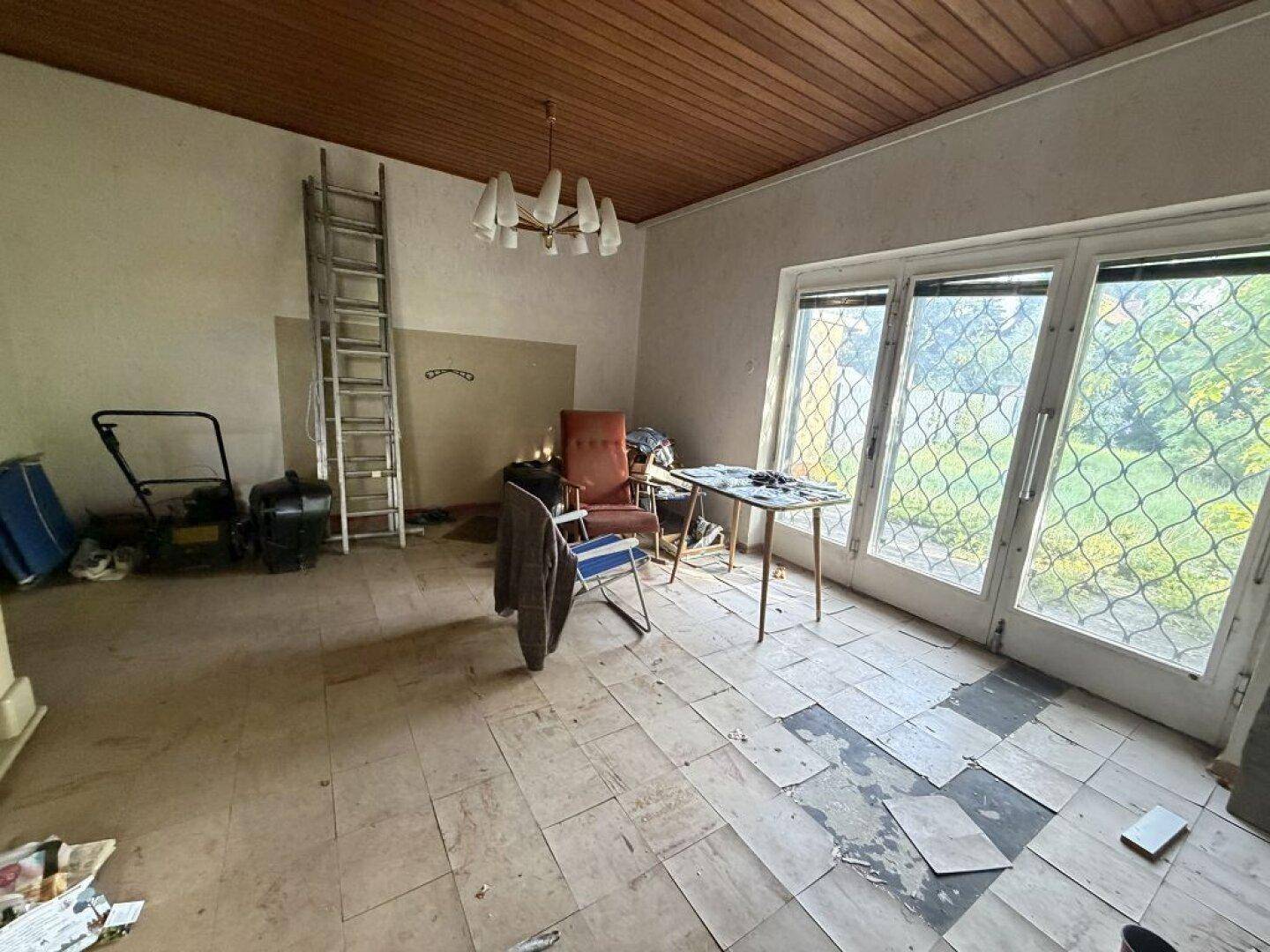 Renovierungsbedürftiger Innenraum mit Holzbalkendecke, Fliesenboden und großen Fenstern zum Garten.