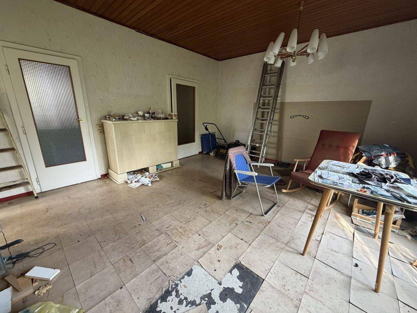 Unaufgeräumter Innenraum mit Holzbalkendecke, beschädigtem Fliesenboden und zwei Türen.