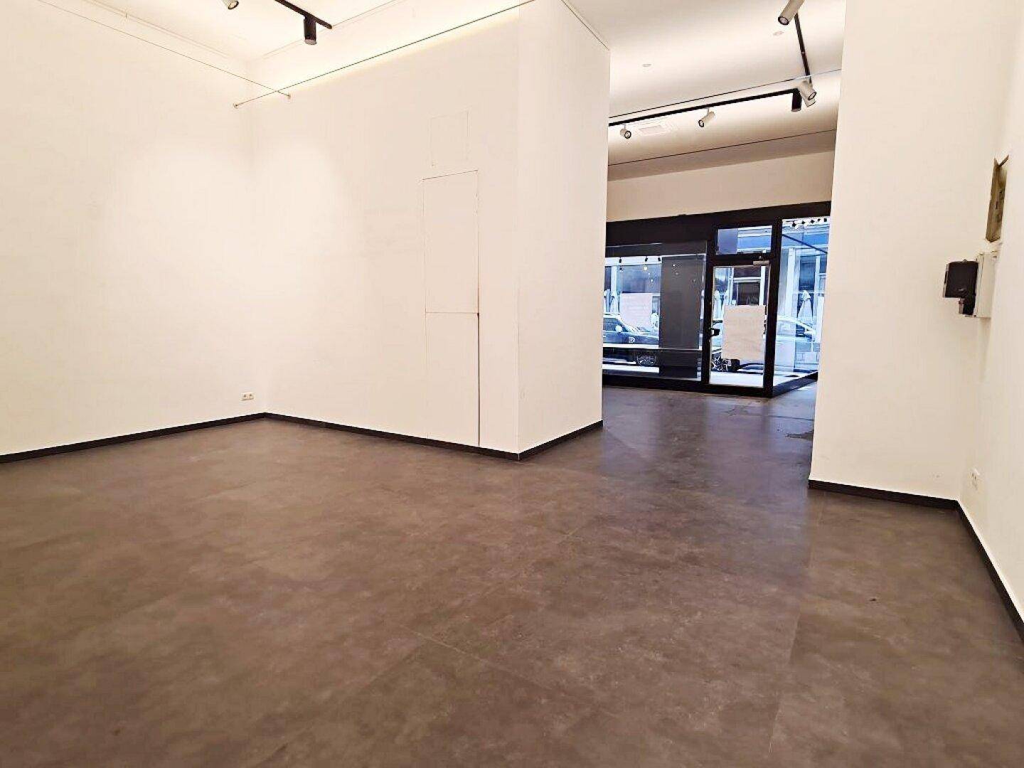 Leerer Geschäftsraum mit großen Schaufenstern im Hintergrund, weißen Wänden und dunklem Boden.