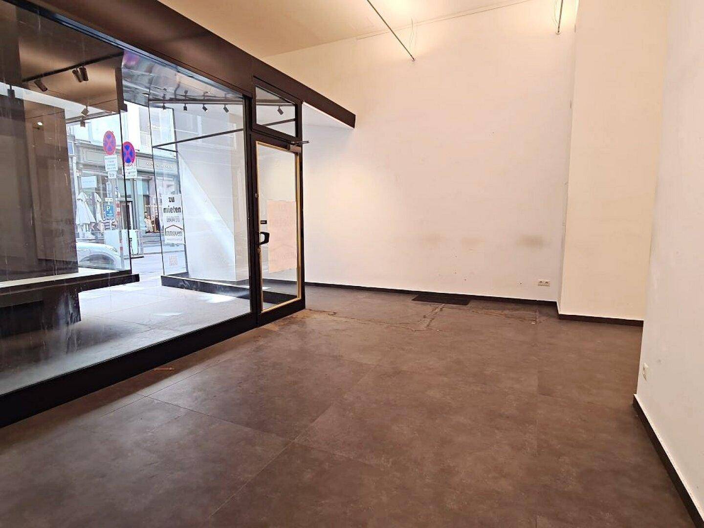 Leerer Ladenbereich mit großen Schaufenstern und Eingangstür, die den Blick auf die Straße freigibt.