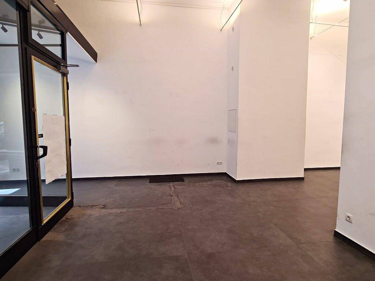 Eingangsbereich eines Geschäftsraums mit dunklem Boden und weißen Wänden, der zur Hauptfläche führt.