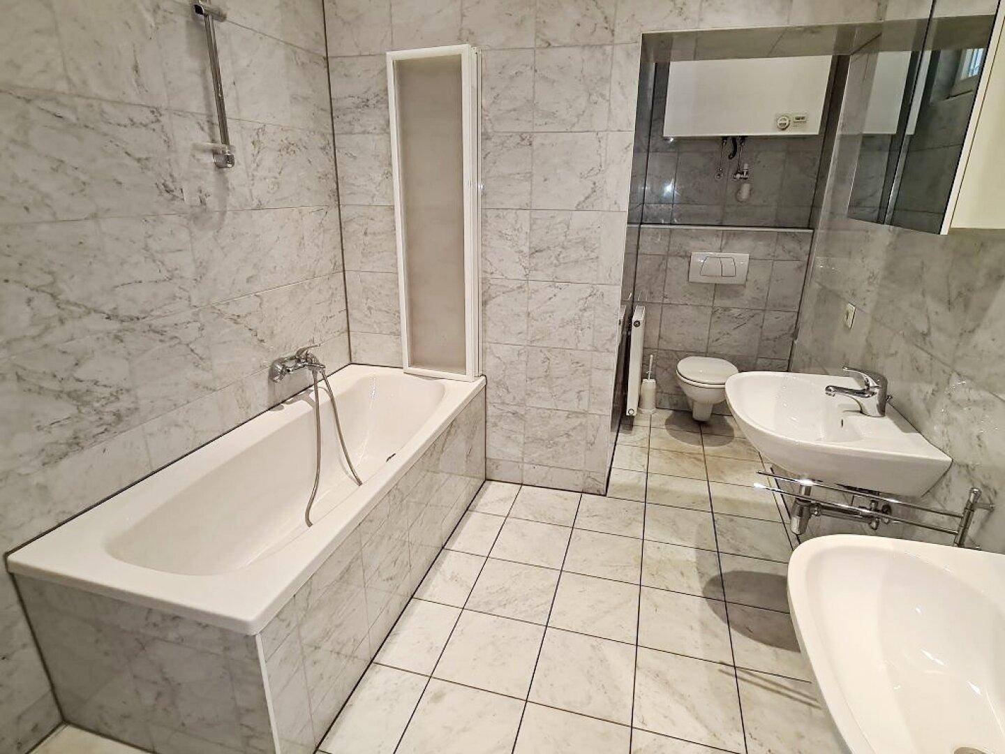 Helles Badezimmer mit Badewanne, Dusche und Waschbecken, komplett gefliest in Marmoroptik.