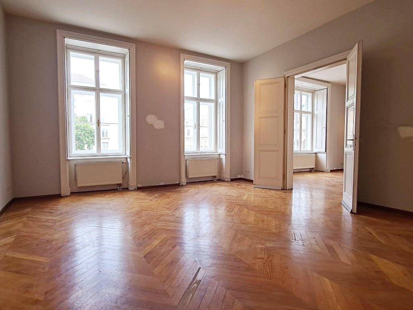 Helles Zimmer mit Fischgrätparkettboden, hohen Fenstern und Zugang zu einem weiteren Raum.