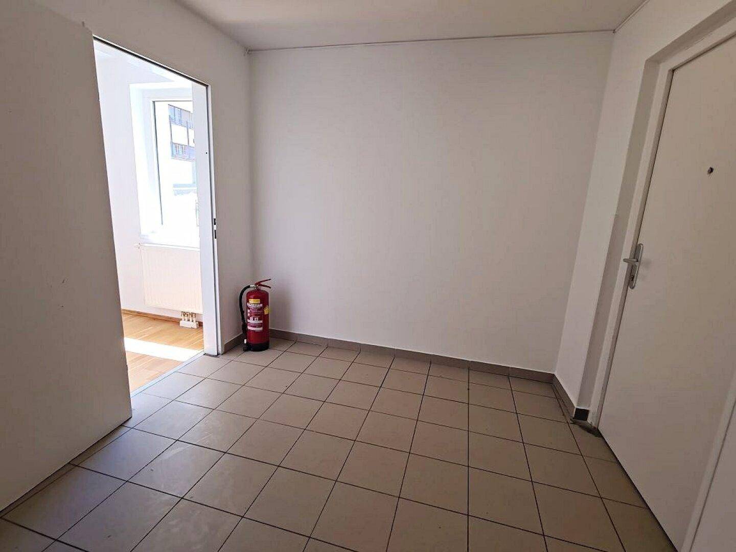 Heller Flur mit Fliesenboden und Zugang zu einem Zimmer mit Parkett, inklusive Feuerlöscher.