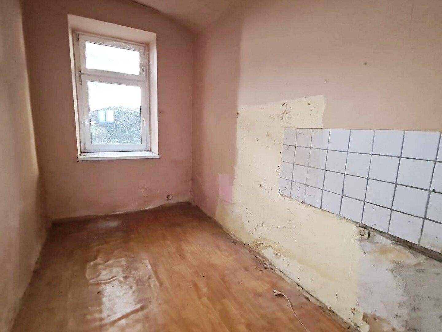 Renovierungsbedürftige Küche mit Fenster, weißen Wandfliesen und beschädigtem Holzboden.