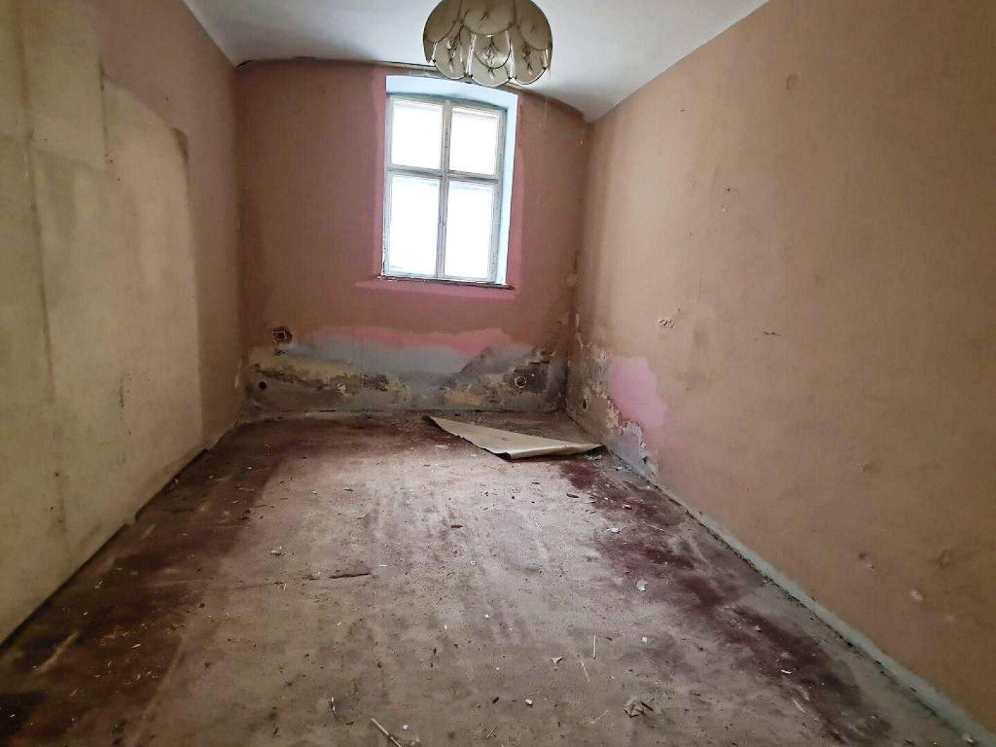 Sanierungsbedürftiges Zimmer mit Fenster, alter Deckenlampe und abgenutztem Bodenbelag.