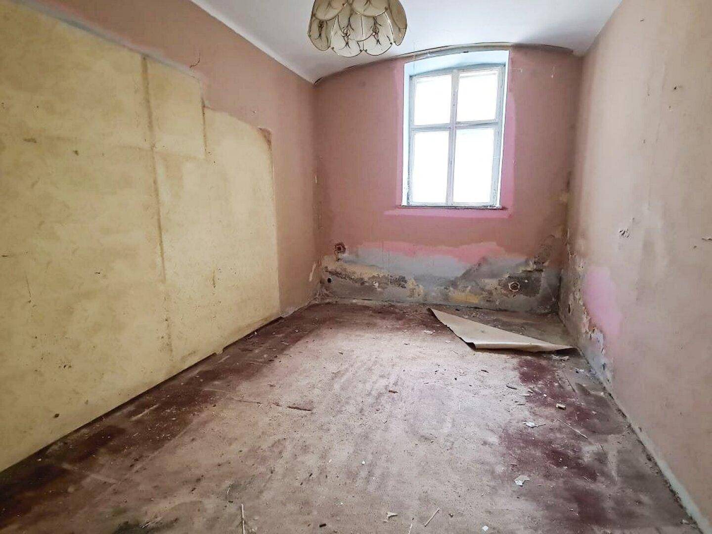 Kleines Zimmer in sanierungsbedürftigem Zustand mit Fenster und alter Deckenleuchte.