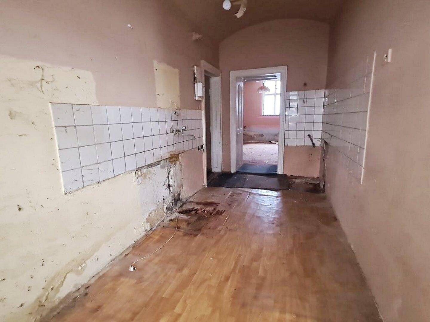 Küche in schlechtem Zustand mit Fliesen und Holzboden, Blick in angrenzendes Zimmer.