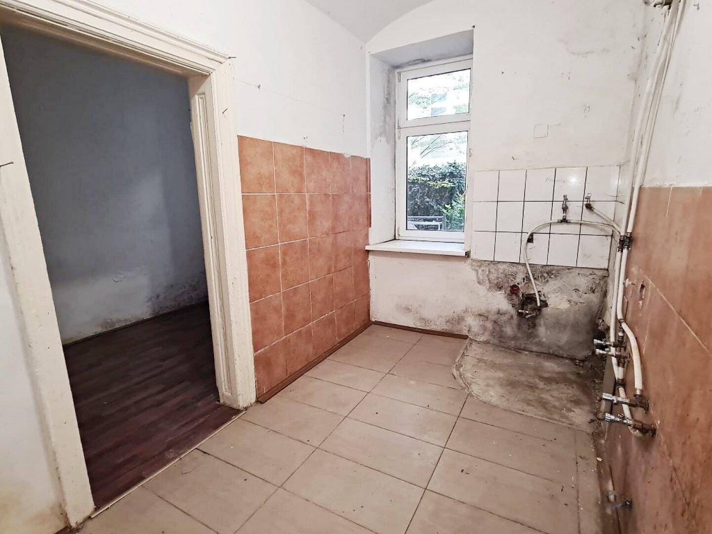 Küche mit braunen Wand- und Bodenfliesen, Fenster und sichtbaren Wasserleitungen.