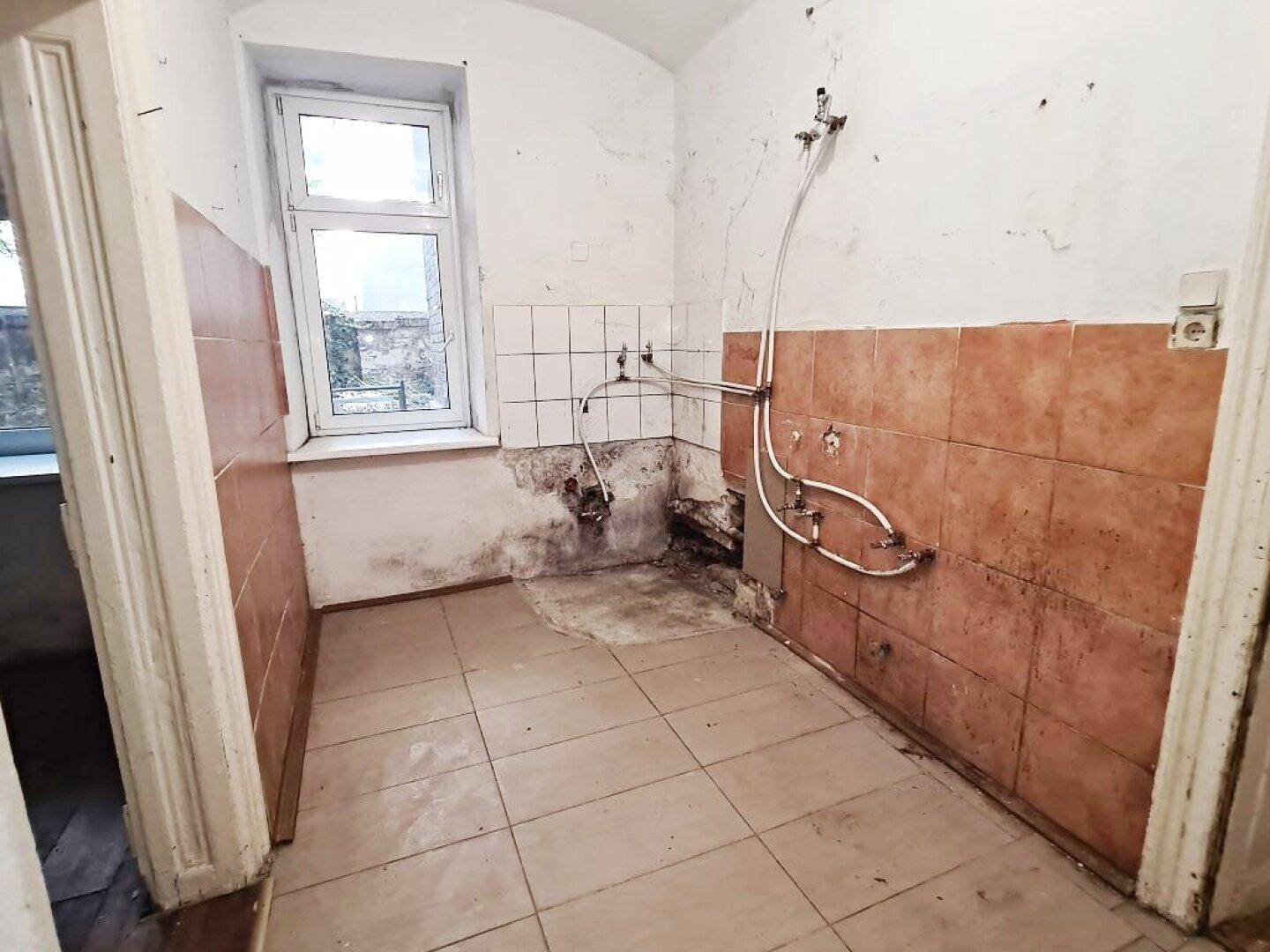 Renovierungsbedürftige Küche mit braunen Fliesen, Fenster und freiliegenden Wasserinstallationen.